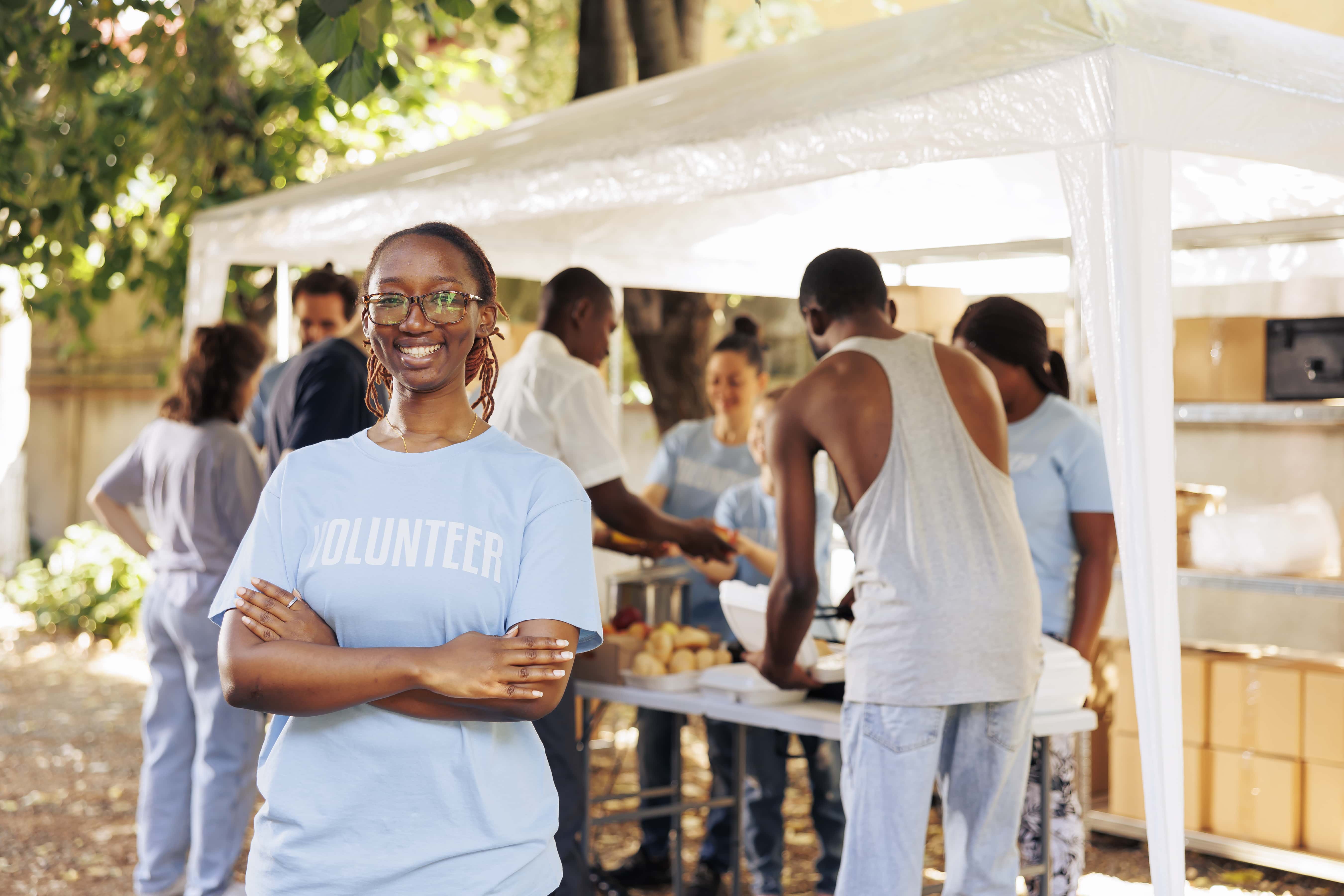 young-black-woman-with-glasses-stands-outdoors-arms-crossed-looking-camera-diverse-group-volunteers-supports-non-profit-program-dedicated-hunger-relief-helping-needy